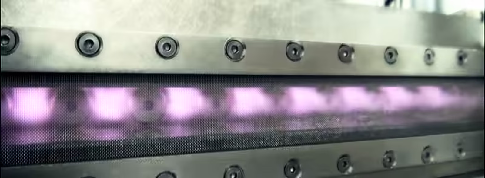 A row of magenta plasma discharges glowing between the electrode bars inside the chamber of an SCI QML inline plasma system